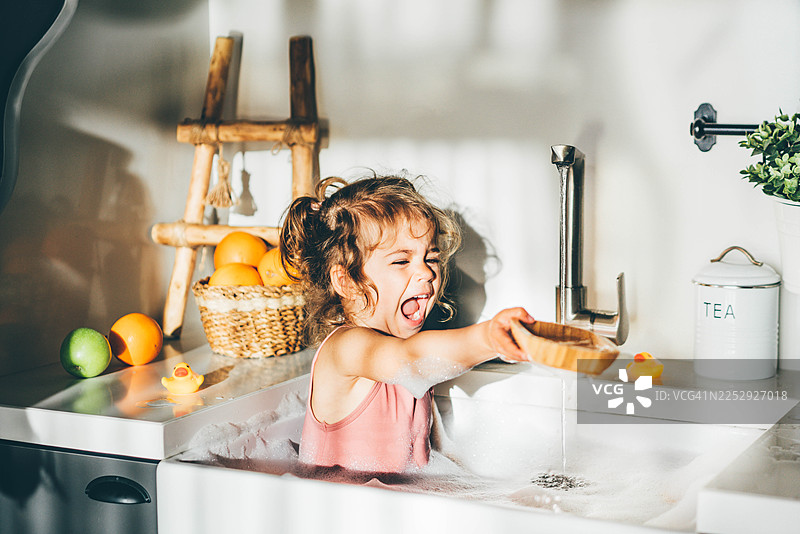 一个快乐的小女孩坐在装满泡沫的厨房水槽里，开心地笑着。图片素材