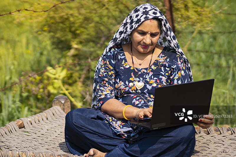白天，一位中年印度妇女和男子坐在农场附近的木床上，使用笔记本电脑。图片素材