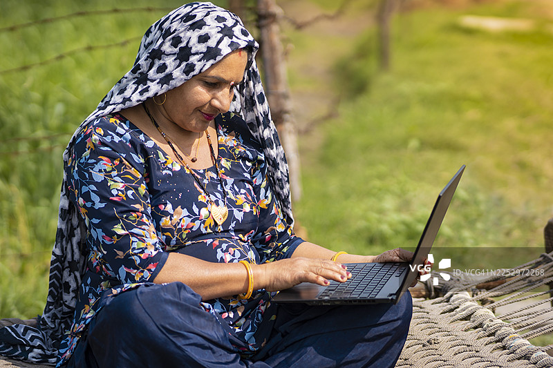 白天，一位中年印度农妇和一名男子坐在小麦农场旁的木床上使用笔记本电脑。图片素材