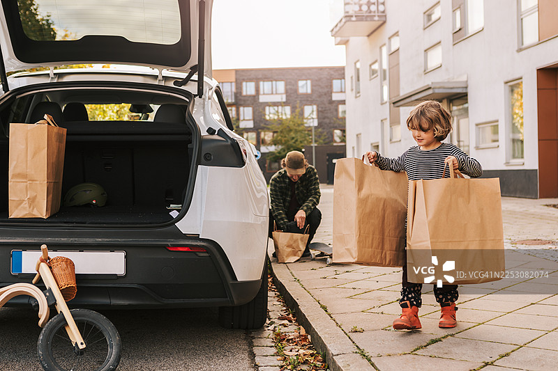 小男孩在帮爸爸把购物袋装进汽车后备箱图片素材