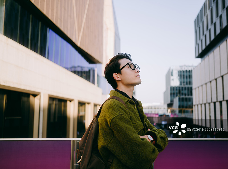 一位自信的年轻亚裔商人抱着双臂，站在现代办公楼前。图片素材