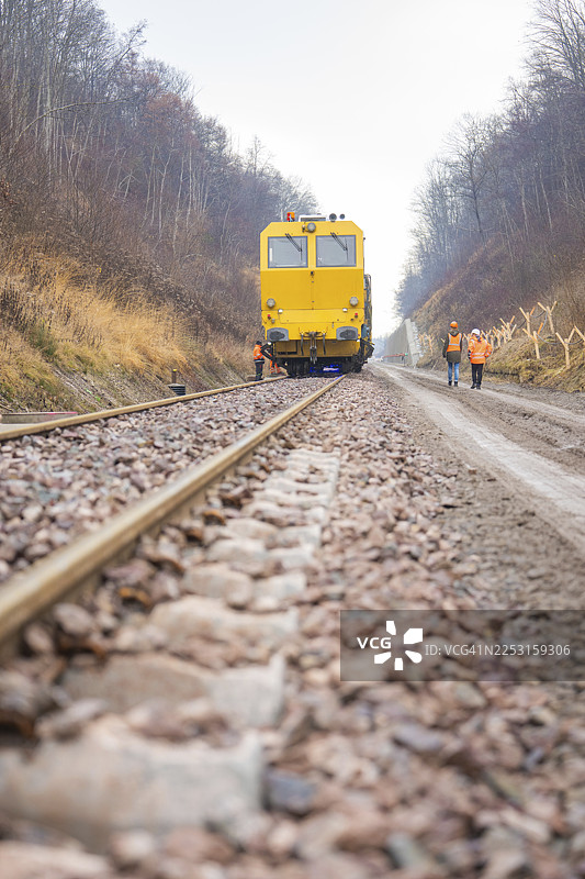 一辆黄色的工程火车在铁轨上行驶，铁轨上有工人在进行填土作业，地点位于德国赫尔曼·黑塞的阿尔滕施泰特，卡尔夫县。图片素材