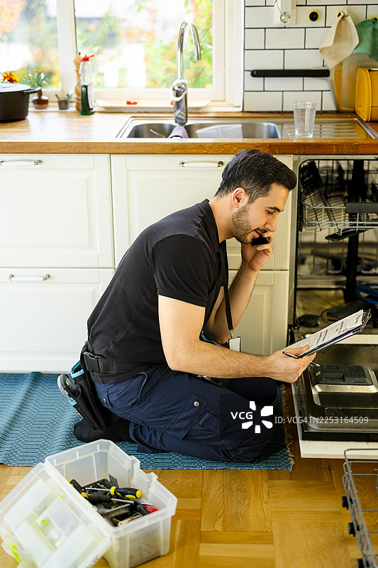 男性技术人员在家中洗碗机旁，一边看表格一边用智能手机打电话。图片素材