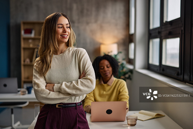 自信的女性双手抱臂站立在现代办公室，旁边有同事正在使用笔记本电脑图片素材