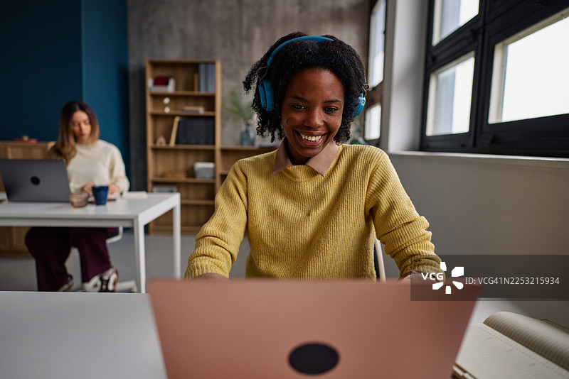 一位年轻女子戴着耳机，在明亮现代的办公室里使用笔记本电脑，她面带微笑，神情专注。图片素材