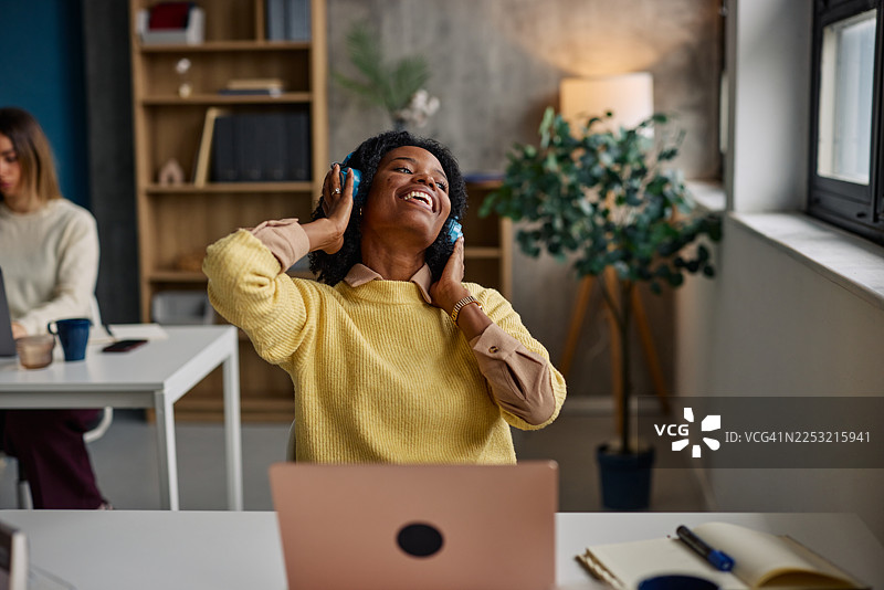 穿着黄色毛衣的快乐女子在现代办公室桌前戴着耳机享受音乐图片素材