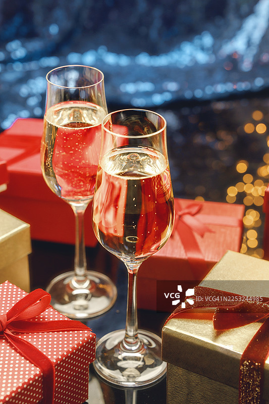 两个香槟酒杯和优雅的金色礼品盒，配有红色天鹅绒蝴蝶结，置于节日的蓝色背景上，背景中点缀着金色串灯和闪光散景效果，供模型参考。图片素材