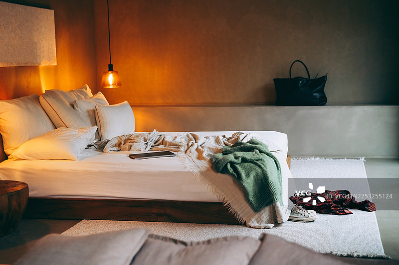 现代风格的舒适温馨卧室，床铺凌乱，灯光柔和，有绿色毛衣、运动鞋、笔记本电脑和散落在地上的衣物。图片素材