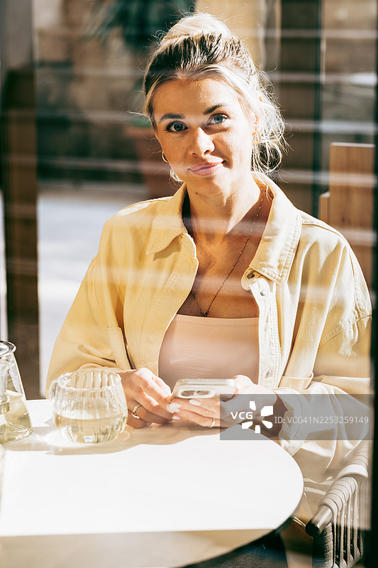 一位自信的年轻女子坐在咖啡馆的桌子旁，从手机上抬起头，带着一丝微笑，透过阳光明媚的窗户捕捉到。图片素材