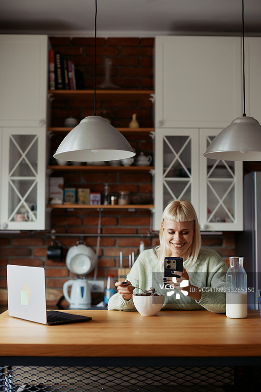 一位年轻的时尚女性在舒适的厨房里享受她的晨间例行公事，吃早餐时拿着手机。图片素材