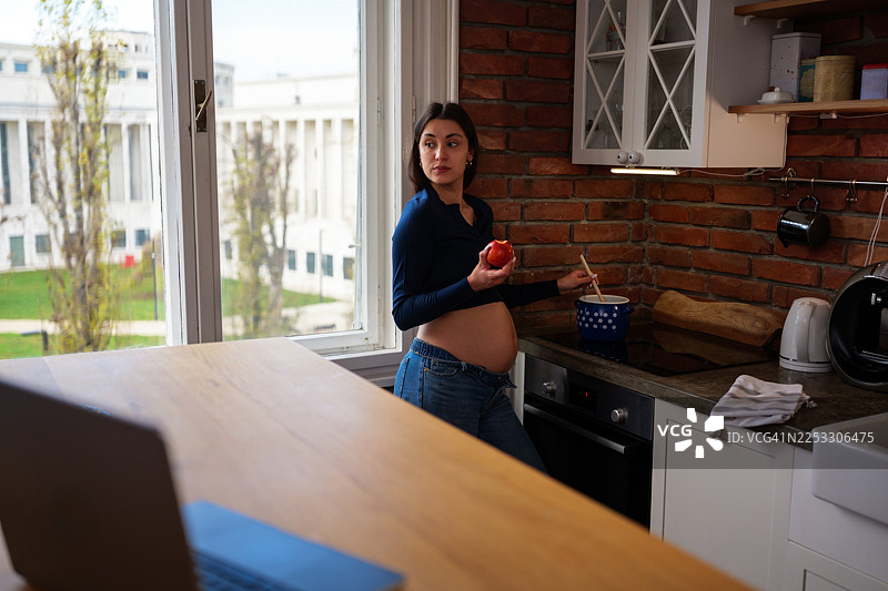 一位年轻的孕妇穿着牛仔裤，站在现代公寓的厨房里，一边搅拌着锅里的食物，一边咬着苹果，她放松而若有所思地为未来的家庭准备健康的餐食。图片素材