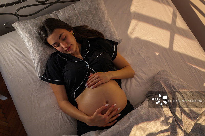 孕妇安详地躺在床上，双手温柔地抚摸着腹部，温暖的晨光透过窗户洒入，凸显了母性的光辉和宁静的期待。图片素材