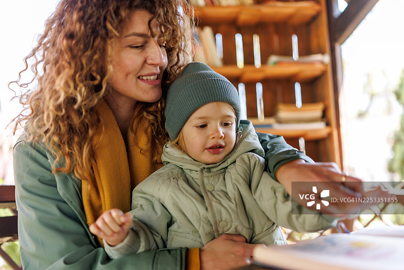 一位微笑的母亲和她可爱的女儿在公共图书馆户外阅读儿童读物图片素材