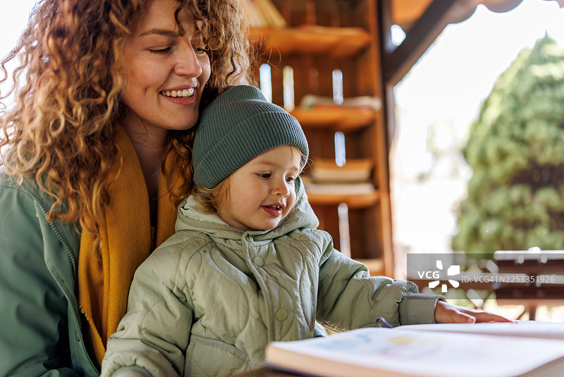 微笑着的母亲和可爱的女儿在城市花园图书馆阅读儿童读物图片素材