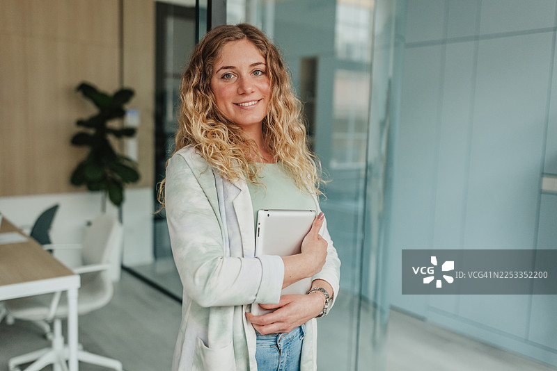 办公室内，一位女士在工作日期间，手持平板电脑，面带微笑地站在玻璃隔板旁。图片素材