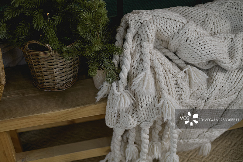 一条带有流苏的白色粗针织毯子，搭在一个木凳上，旁边是盆栽的常青植物和一个编织篮。图片素材