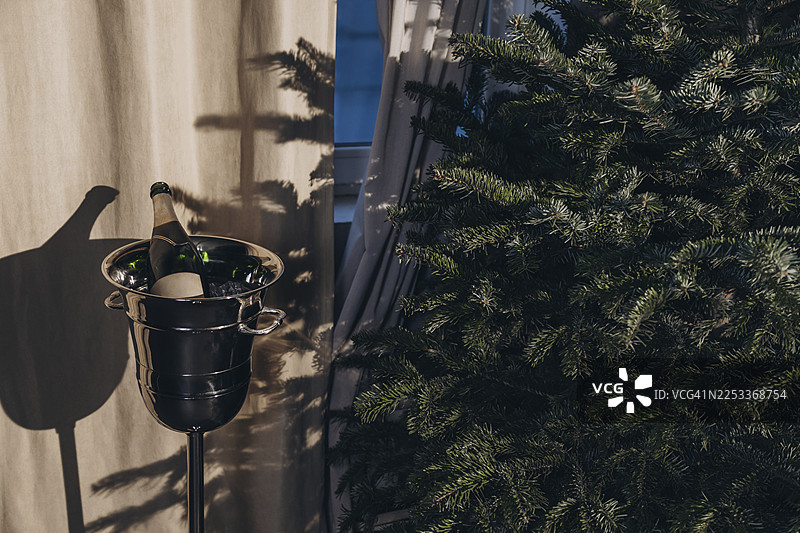 一个银色的香槟桶里装着一瓶酒和冰块，旁边是圣诞树，在浅色幕布上投下阴影。图片素材