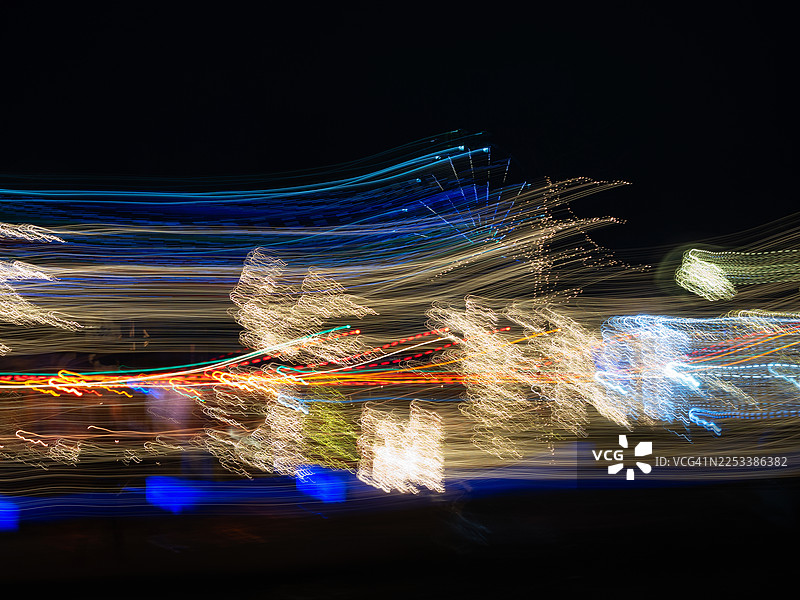 抽象的城市夜景，刻意移动的相机，用暖白色灯串和蓝色灯光装饰的树木。白色、黄色、蓝色、红色和绿色的水平光轨横跨画面，背景为深色。图片素材