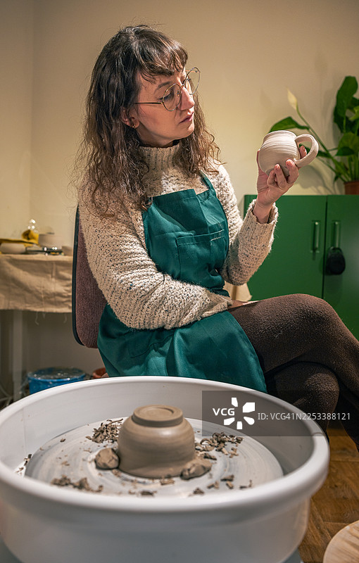 一位时髦女士坐在陶轮旁修整碗，同时审视着一个手工陶瓷杯。图片素材