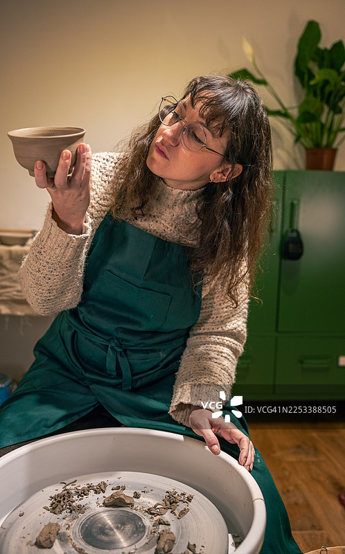 一位时尚女性坐在陶轮前，审视着手工制作的陶碗。图片素材