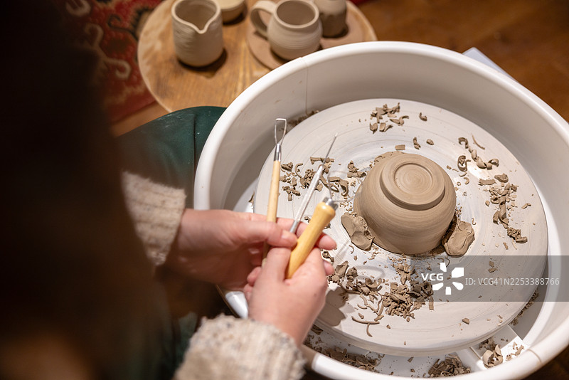 一位女士坐在陶轮上，用锋利的工具修整手工陶碗。图片素材