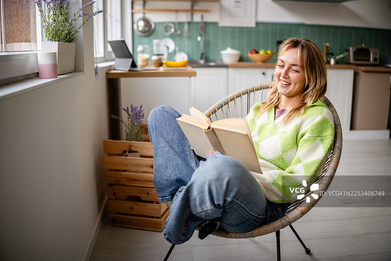 一位年轻女子在家中阳光明媚的日子里，舒适地坐在椅子上，在温馨的厨房里享受阅读的时光图片素材