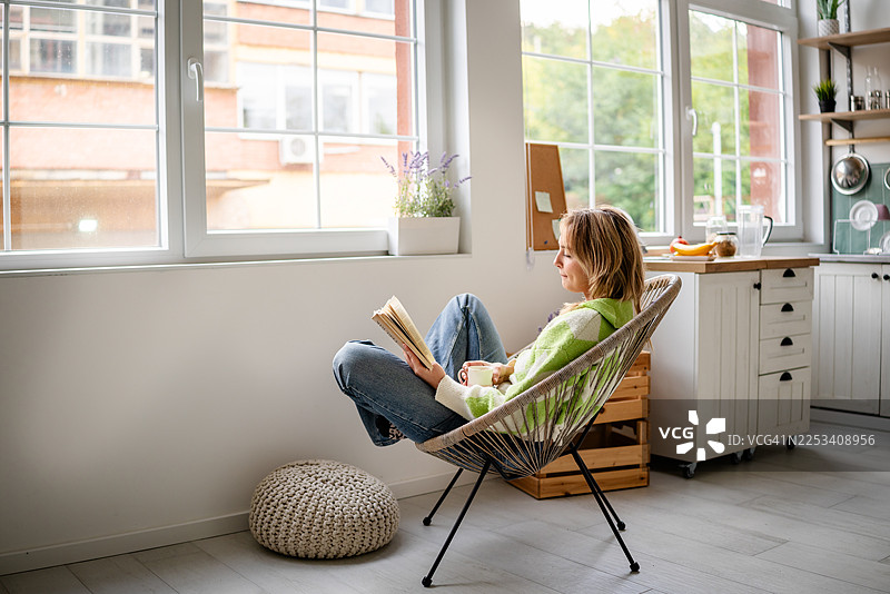 年轻女子在公寓里明亮的厨房里享受午后阅读时光，自然光倾泻而入。图片素材