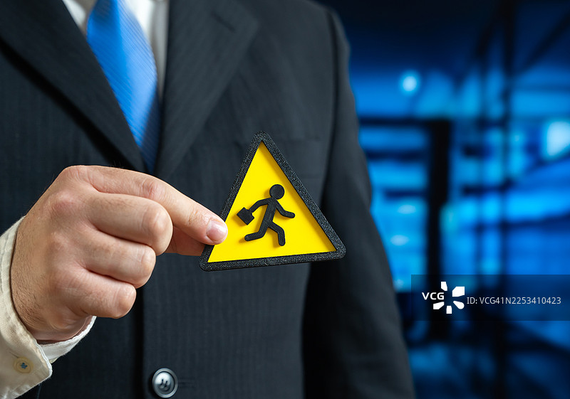 一名商界人士举着一个黄色警告标志，上面描绘了一个带着公文包奔跑的人，象征着潜在的商业风险或快速撤离图片素材