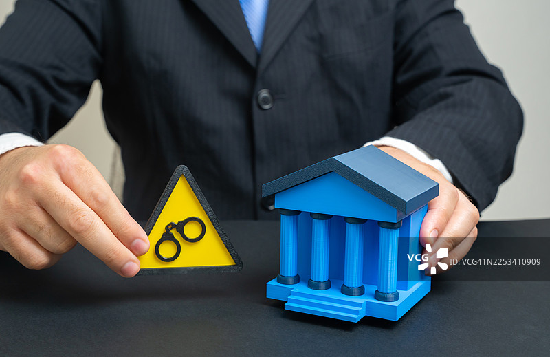 一名商人手持警告标志和一副手铐，旁边有一个银行建筑模型，象征着金融领域的法律风险图片素材