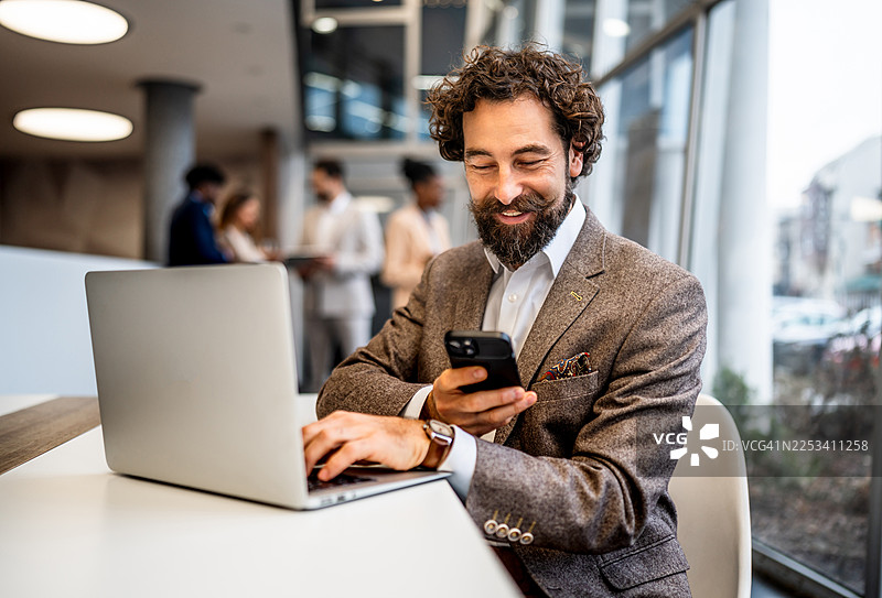 一位面带微笑的商人在现代办公室里同时使用笔记本电脑和智能手机，处理多项事务图片素材