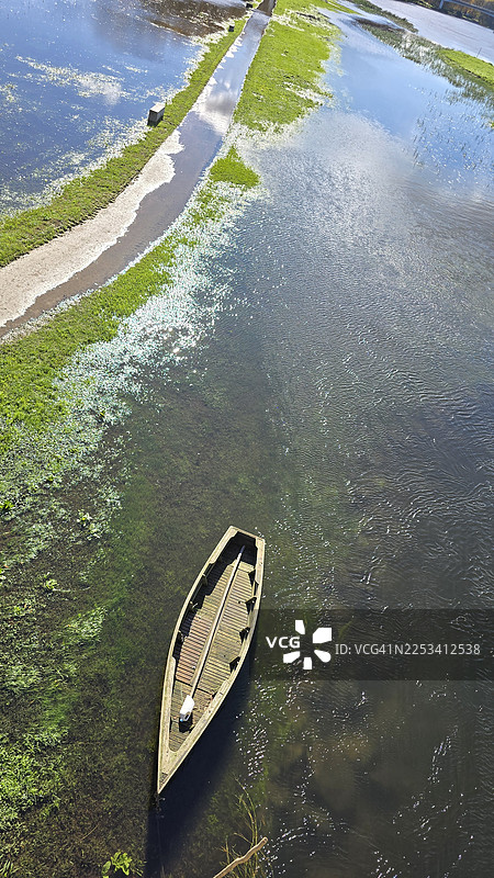 从高处俯瞰，一艘木制划艇停泊在河边，河岸绿意盎然，天空阳光明媚。图片素材