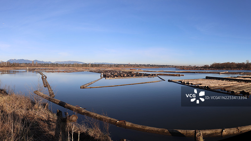 广阔河流上的木材集材带的全景，远处是山脉，天空湛蓝，不列颠哥伦比亚省列治文市菲沙河畔的住宿加早餐旅馆图片素材