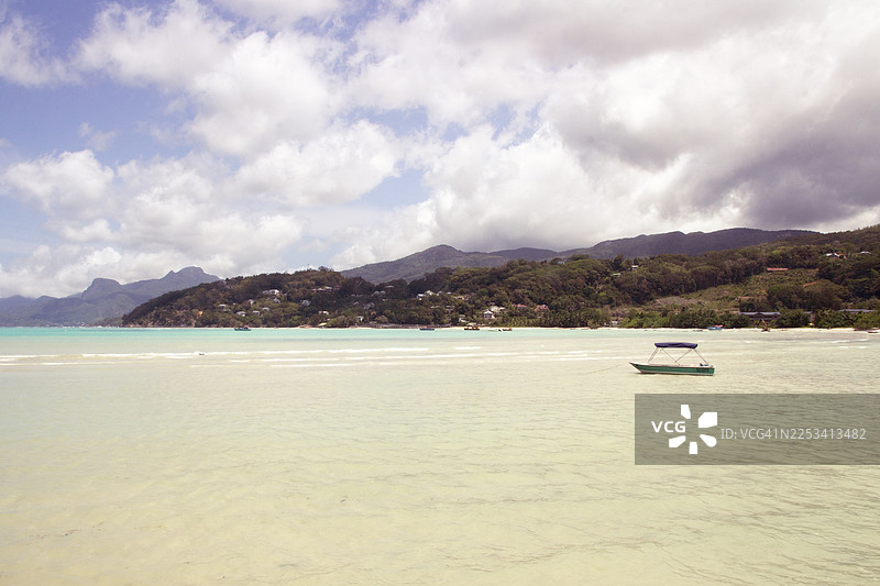 宁静的热带海景，一艘小船漂浮在清澈的绿松石色海面上，背景是郁郁葱葱的绿色山脉，天空多云，塞舌尔，格兰安塞马埃图片素材
