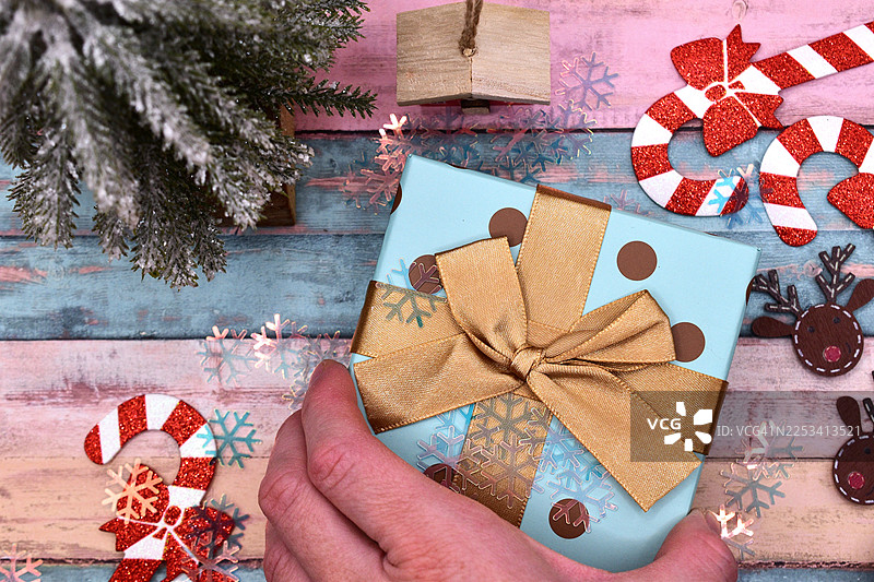 一只手拿着浅蓝色礼品盒，上面系着金色丝带，背景是乡村木质桌面，装饰着节日圣诞饰品图片素材