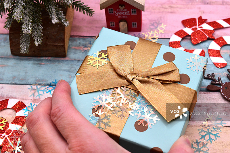 裁剪的手，手里拿着一个带有金色丝带和雪花纸屑的蓝色圣诞礼物，周围是木质背景上的节日装饰图片素材