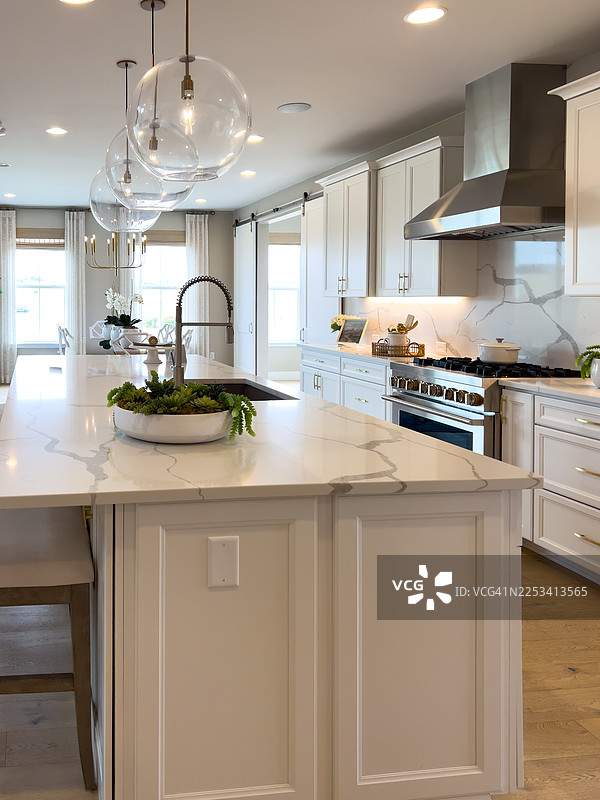 现代豪华厨房内部，配有白色大理石台面和不锈钢电器，设有一个带水槽的大型岛台和透明玻璃吊灯图片素材