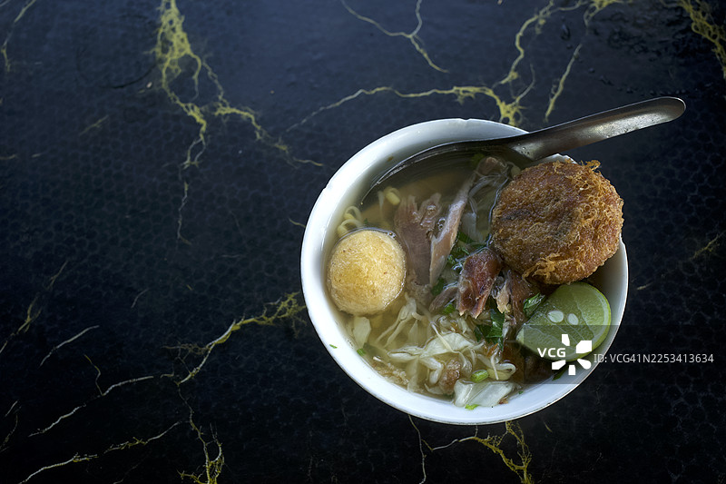 俯视角度拍摄的印尼传统 Soto 汤，配有青柠和炸配料，放在深色图案的桌子上图片素材