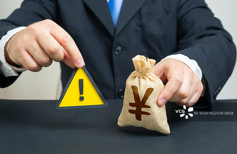 一名商人拿着一个日元符号的钱袋和一个警告标志，象征着财务风险和投资谨慎图片素材