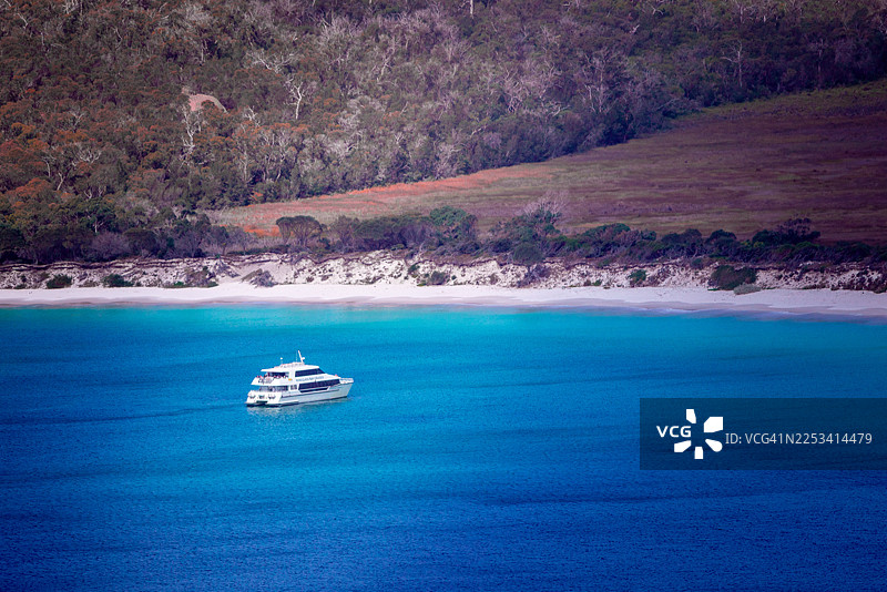 从高处俯瞰一艘小型游轮在澳大利亚的葡萄酒杯湾瞭望点附近，驶过绿松石般的海面，海边是白色沙滩和森林海岸线图片素材