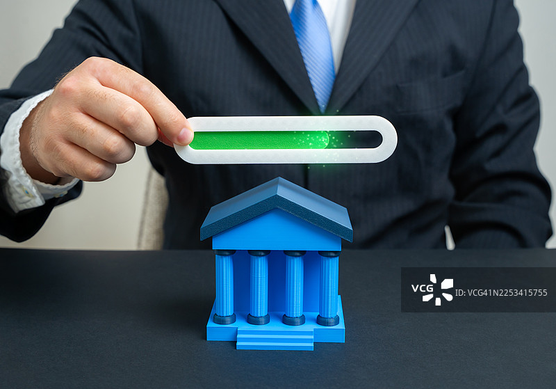 商人举着发光的进度条，象征着财务增长或项目完成，进度条上方是一个模型银行建筑，摆放在办公桌上图片素材
