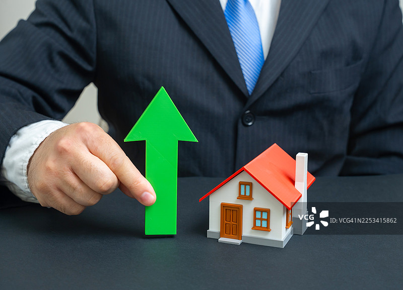 一位商人手持指向上的绿色箭头，旁边有一个微型房屋，象征着房地产的增长图片素材