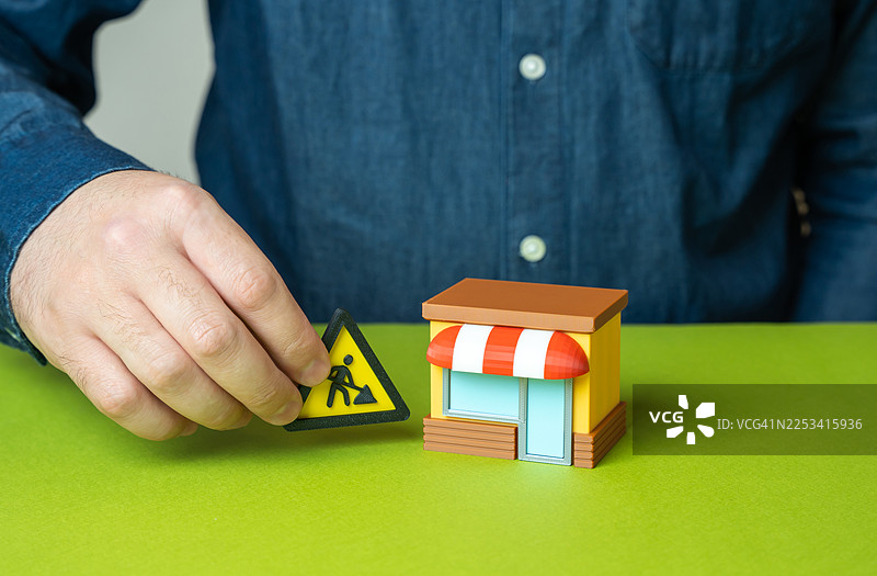 一只男人的手将一个“施工中”的标志放在一个迷你商店模型旁边，象征着商业发展或建设图片素材