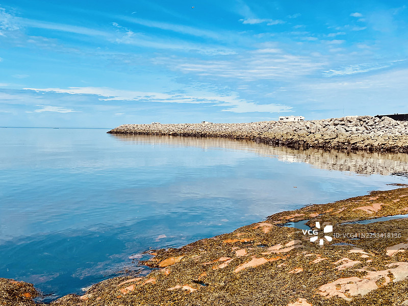 宁静的海滨风光，拥有平静的蓝色大海、岩石防波堤以及海藻覆盖的海岸，天空晴朗图片素材