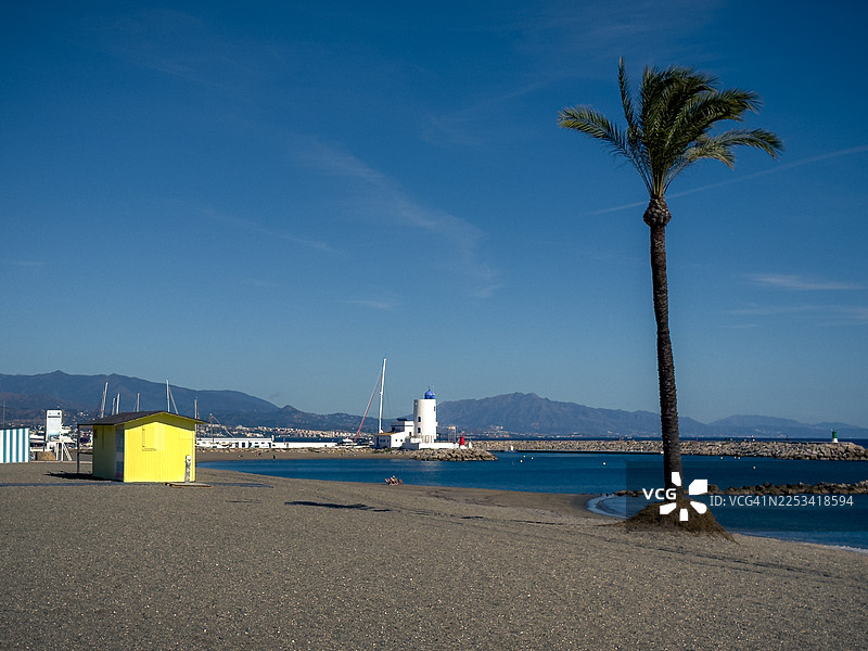西班牙马尼尔瓦，杜克萨，晴朗蓝天下有棕榈树、灯塔和山脉的宁静海滩风景图片素材