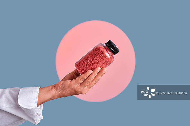 蓝色背景上粉色圆圈映衬下，手持红色颗粒瓶子的特写图片素材