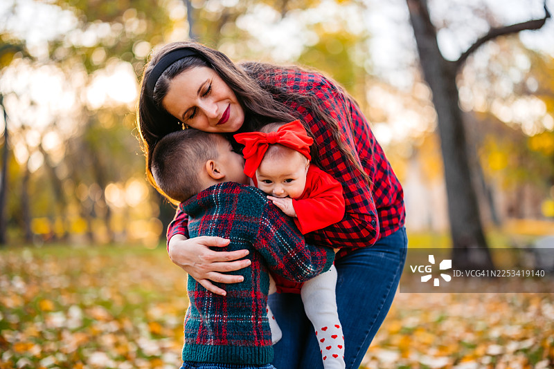 秋日里，年轻的母亲在公园里拥抱着她学龄前的小儿子和年幼的女儿。图片素材