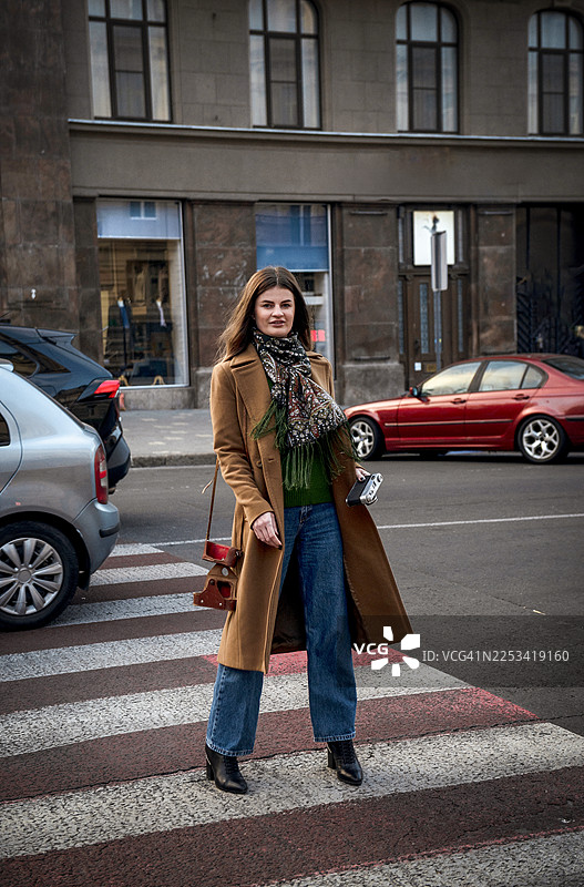 身穿长款外套和牛仔裤的时尚女子站在城市街道上，手持相机图片素材