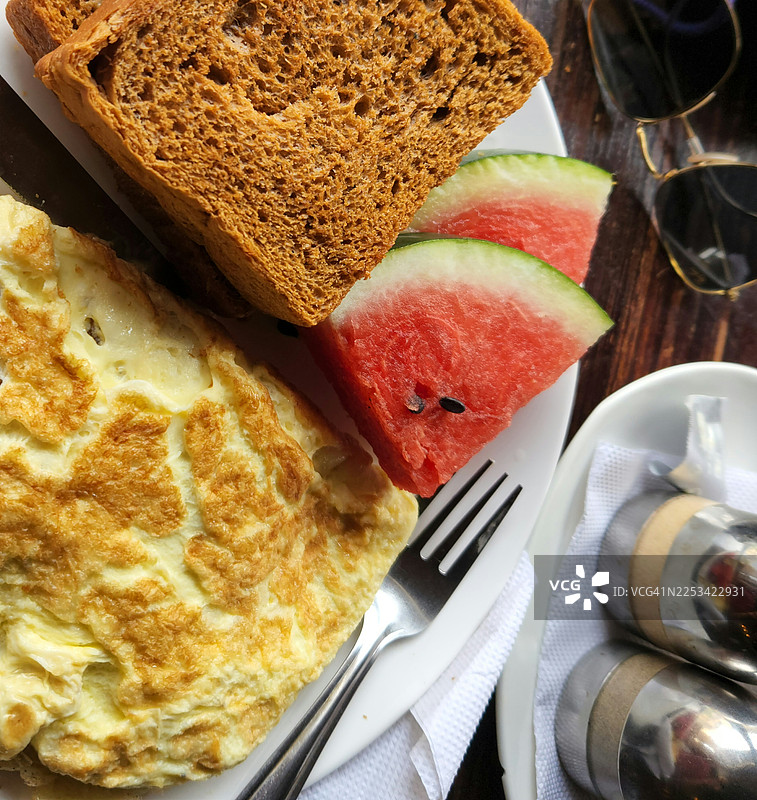 高角度俯视画面，展示了木桌上丰盛美味的早餐，包括煎蛋卷、吐司和新鲜西瓜片，旁边还放着一副太阳镜。图片素材