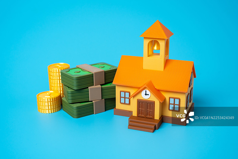 蓝色背景上的教学楼模型，堆放着钱币和硬币，象征着教育成本和资金图片素材