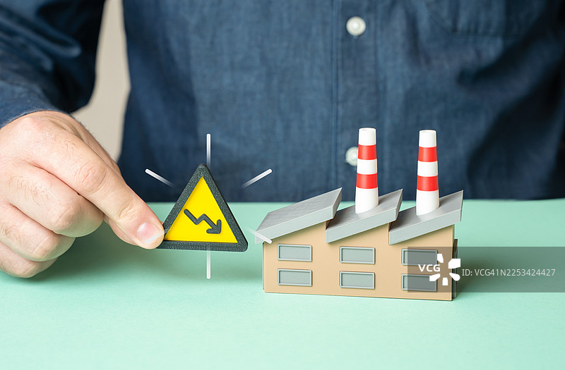 一只男人的手将带有向下箭头的警告标志放在一个模型工厂旁边，象征着工业衰退或经济衰退图片素材